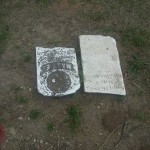 Two tombstones are resting on the ground, with a natural setting around them, suggesting a peaceful memorial area.